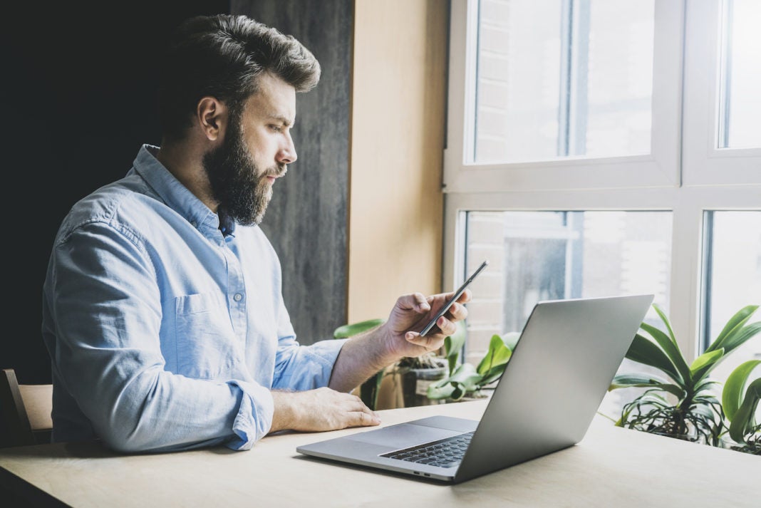 Image of business person sitting at desk with laptop and plants, looking at smartphone.