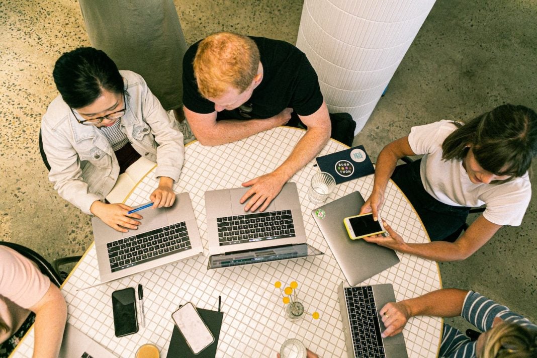 Image of business professionals working on laptops, sitting around a circular table. Meant to represent a brainstorm session for low-budget marketing and advertising ideas.