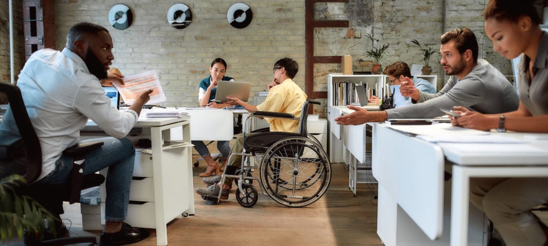 Portrait of young male worker in wheelchair talking to female colleague while presenting ideas using laptop in co-working space.