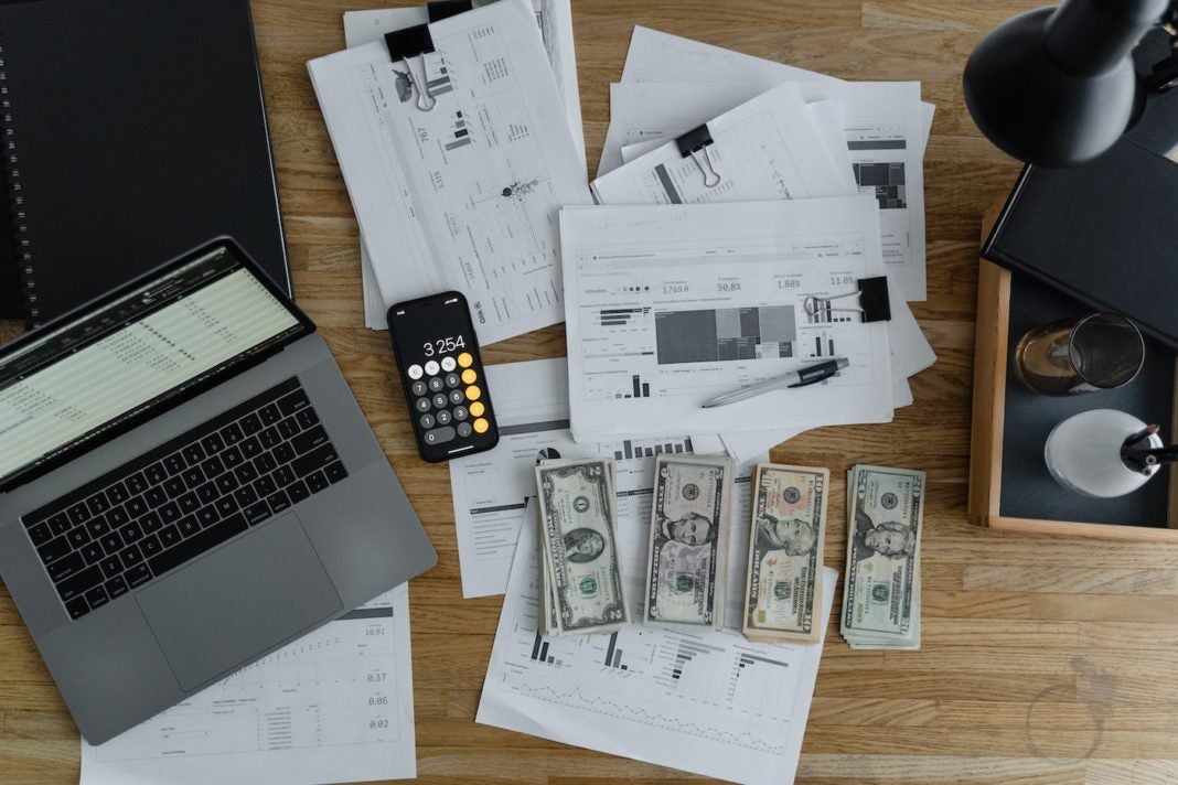 Photo of payroll software on a laptop with money, reports, and a calculator spread out on the table.