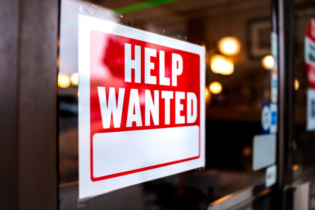 Sign text closeup for help wanted with red and white colors by entrance to store shop business building during corona virus covid 19 pandemic.